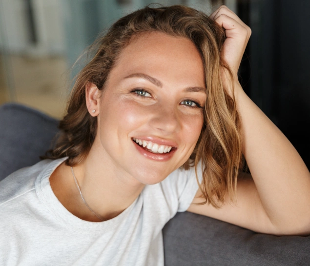 Young woman smiling brightly while touching her hair, wearing white shirt indoors.