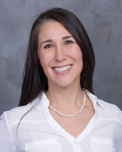 Professional headshot of female dentist smiling, wearing white coat and pearl necklace against gray background.