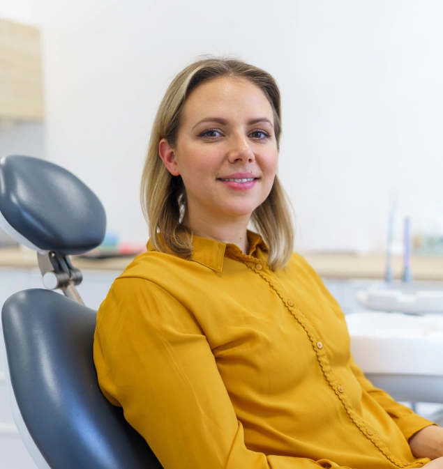 Woman in yellow shirt smiling in dental office chair with window view behind her.