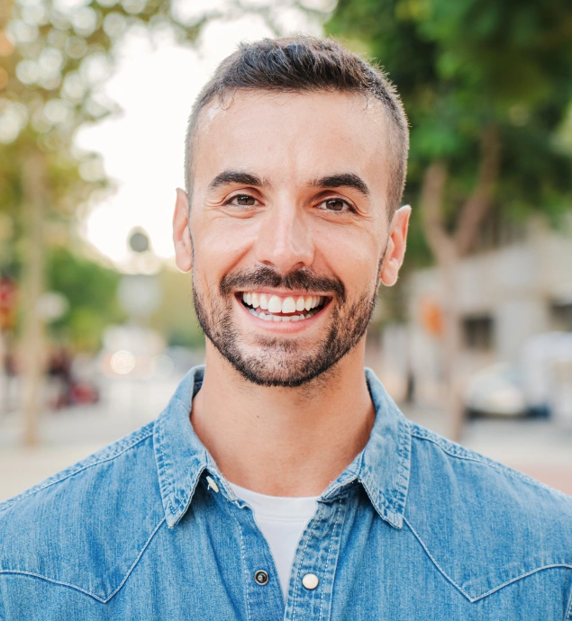 Smiling man in denim shirt poses for professional headshot outdoors with blurred background.