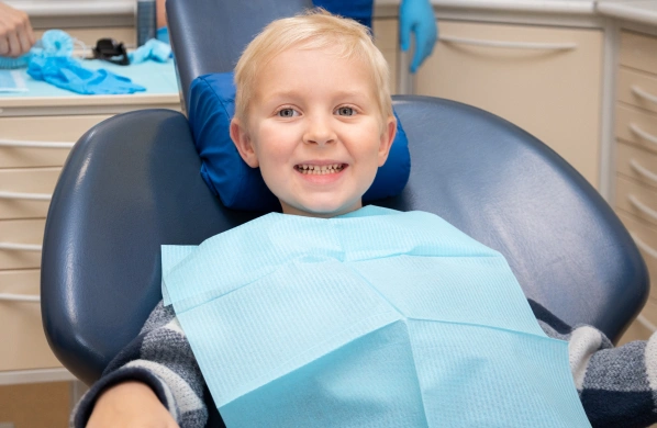 Young child smiling in dental chair wearing blue bib at dentistry office.