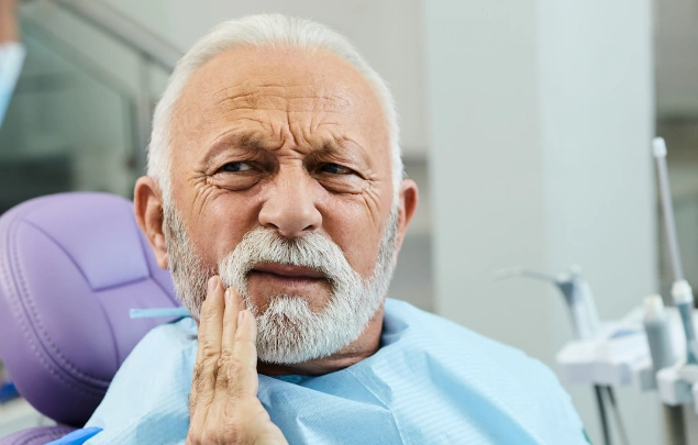 Elderly man with white beard in dental chair touching face, expressing discomfort or concern.