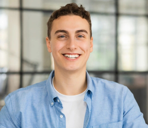 Young man smiling at camera wearing light blue shirt in modern office setting.