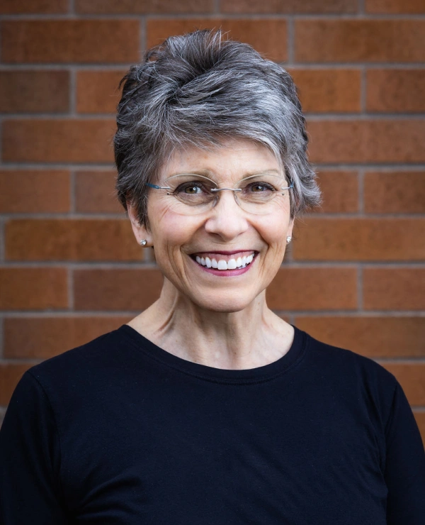 Professional headshot of smiling woman with gray hair against brick wall background.