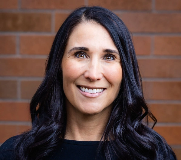 Professional headshot of smiling woman with long dark hair against brick wall background.