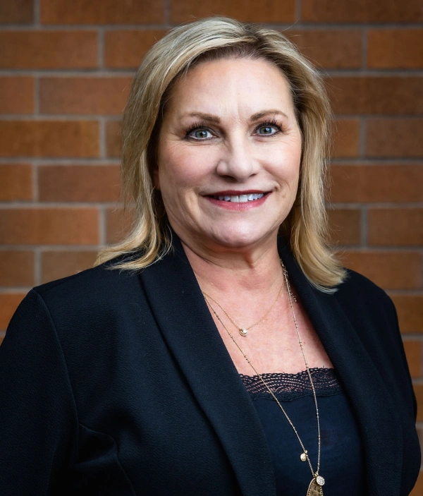 Professional headshot of woman in black blazer against brick wall background.