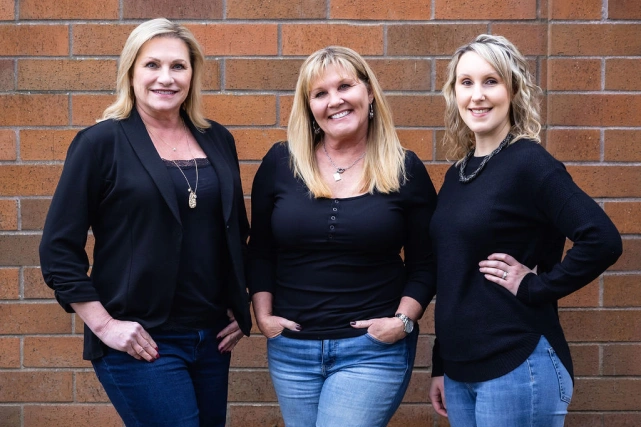 Three women in black tops pose together against a brick wall background.