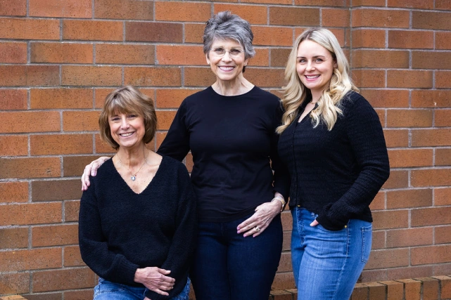 Three women smiling at camera against brick wall background for dental practice portrait.