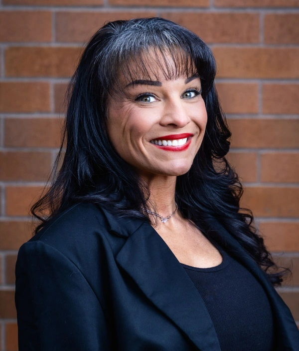 Professional woman in black blazer smiling against brick wall backdrop for dental practice.