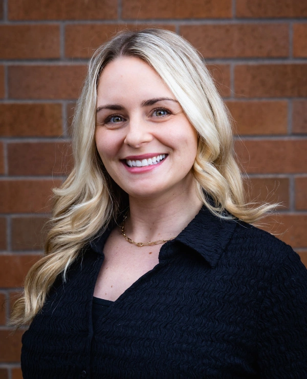 Professional headshot of smiling woman with blonde hair against brick wall background.