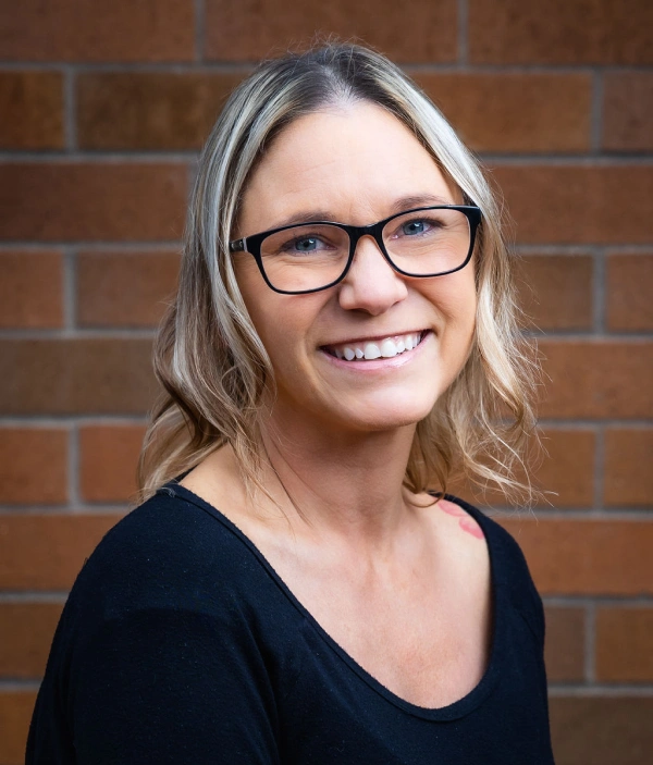 Woman wearing glasses smiling at camera against brick wall background, professional headshot.