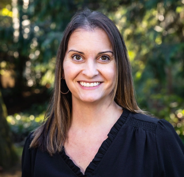 Professional headshot of smiling woman in black top against blurred green foliage background.