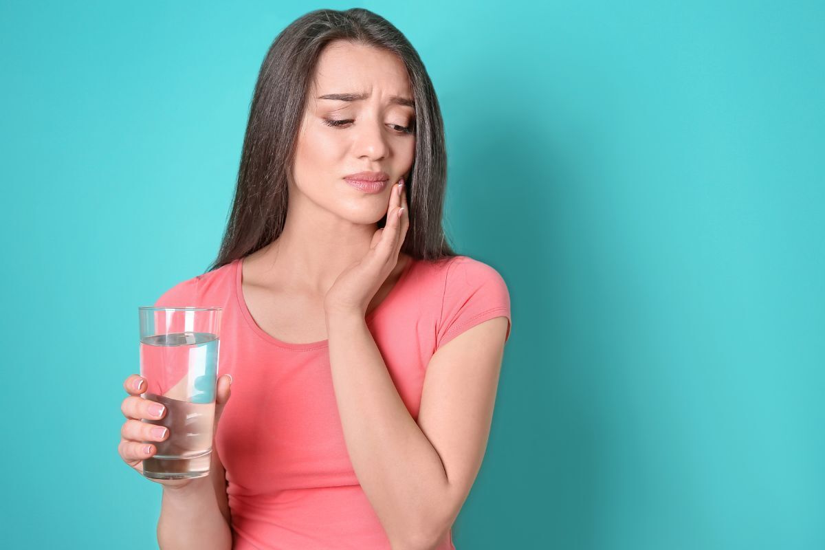Woman in pink shirt holding water glass with hand on cheek, experiencing tooth sensitivity.