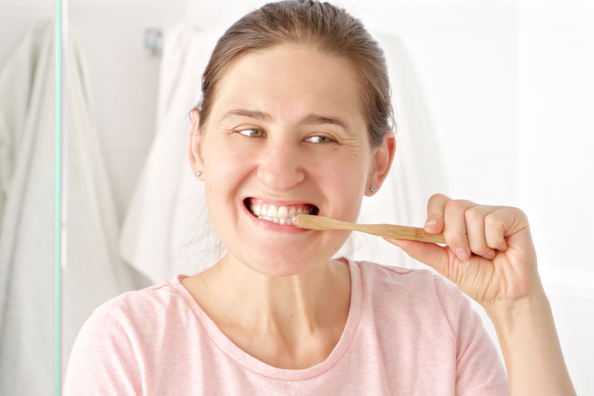 Woman brushing her teeth with a bamboo toothbrush, smiling in a bathroom mirror.